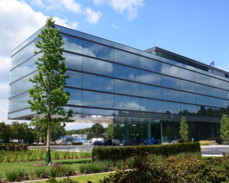 Modernes, verglastes Bürogebäude mit Himmelreflexionen und einem Baum davor.